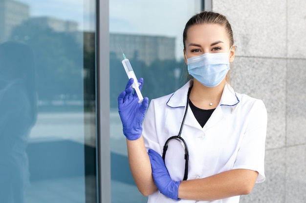 Santé, médecine, profession, concept de personnes - Infirmière orientale avec un stéthoscope portant une blouse médicale, des gants et un masque de protection et tenant une seringue à la main sur le côté avec un espace libre