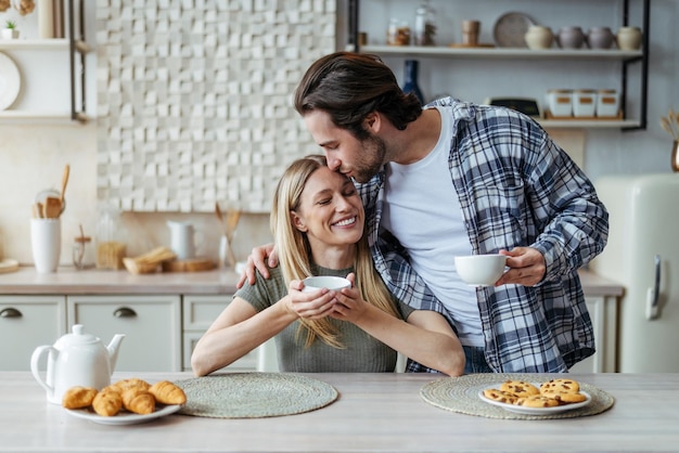 Satisfait jeune homme européen avec chaume bisous femme boire une boisson chaude pendant le temps libre dans le moderne