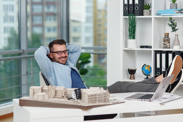 Satisfait de son projet, le designer masculin est assis dans une position détendue, les jambes jetées sur la table dans un studio de design.