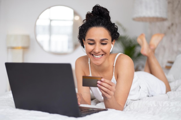 Satisfaite jeune femme brune caucasienne aux cheveux bouclés en pyjama payant de l'argent avec carte de crédit