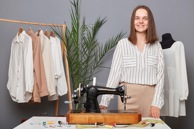 Satisfaite souriante femme aux cheveux bruns égout debout près de sa machine à coudre regardant la caméra étant prête à travailler la couturière sur son lieu de travail