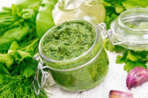 Sauce à l'aneth, persil, basilic, coriandre, autres herbes épicées, ail et huile végétale dans un bocal en verre, gros sel sur fond de planche de bois blanc