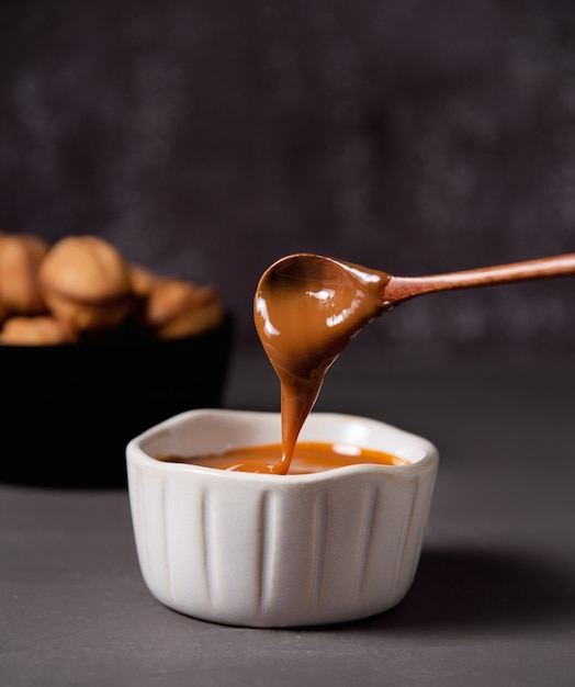 La sauce au caramel maison coule d'une cuillère en bois dans un bol blanc sur fond sombre. Vue de face et gros plan