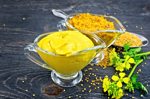 Sauce moutarde et moutarde de Dijon dans deux casseroles en verre, fleur jaune et graines dans une cuillère sur fond de planche de bois noir