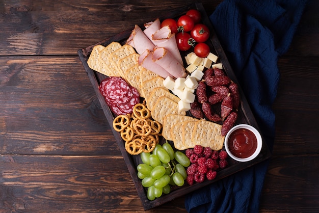 Saucisse, craquelins, fruits et fromage sur planche de charcuterie sur fond de bois foncé, gros plan.