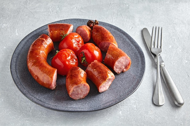Photo saucisse grillée coupée en tranches et tomates frites sur une assiette en céramique grise avec couverts sur une table en béton gris
