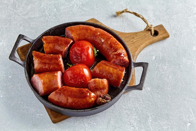 Photo saucisse de porc frite coupée en morceaux et tomates frites sur une poêle en fonte noire sur une planche de bois sur une table en béton gris