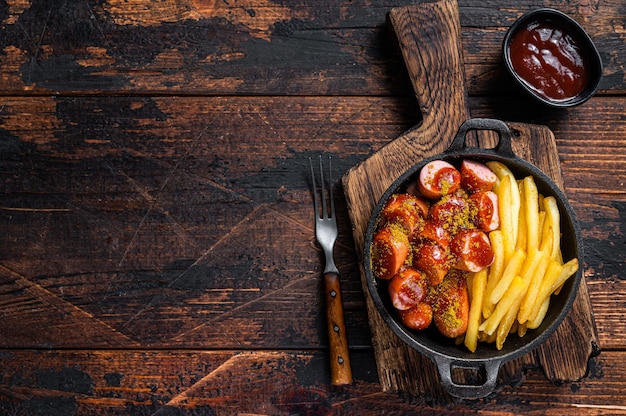 Saucisses au currywurst avec des épices au curry sur des wursts servies des frites dans une poêle. Fond en bois sombre. Vue de dessus. Espace de copie.