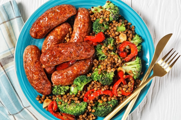 Saucisses de porc italiennes braisées avec des lentilles brunes, du poivron rouge et du brocoli sur une plaque bleue sur un fond en bois blanc avec des couverts dorés, de la moutarde, du thym et du ketchup, vue de dessus, gros plan