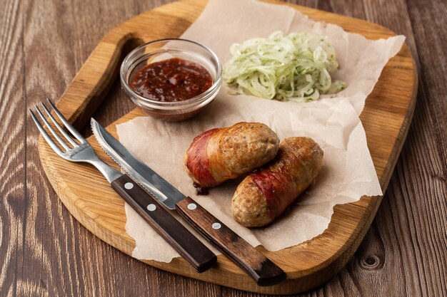 Saucisses de porc prêtes à manger enveloppées de bacon sur planche de bois Saucisses salées frites enveloppées de bacon servies avec oignon et sauce sur fond de bois