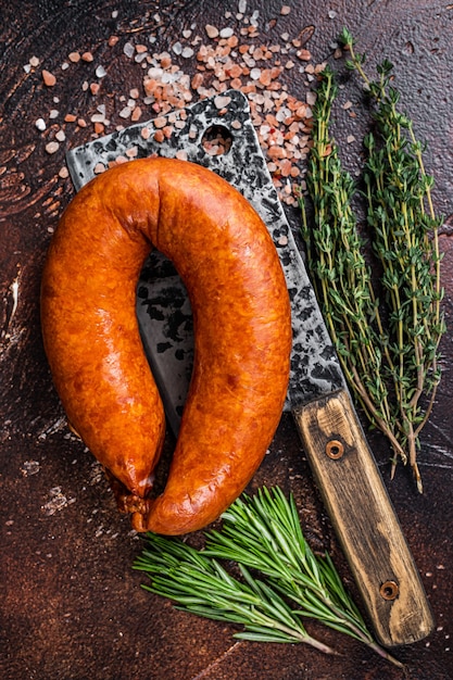 Saucisson fumé à chaud sur un couperet rustique de boucher aux herbes. Fond sombre. Vue de dessus.