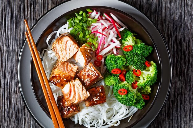 Saumon teriyaki avec nouilles de riz radis brocoli et oignon vert dans un bol avec des baguettes sur une table en bois sombre mise à plat gros plan