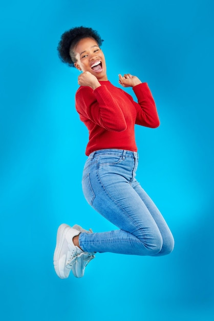 Saut excité et portrait de femme avec un accord gagnant et sourire dans un studio Wow énergie et en l'air avec une personne de sexe féminin et un fond bleu avec une célébration pour l'annonce de la vente et de grandes nouvelles