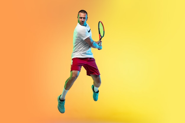 En saut. Un homme de race blanche jouant au tennis sur fond de studio à la lumière du néon. Monter un jeune joueur professionnel masculin en mouvement ou en action pendant un match de sport. Concept de mouvement, sport, mode de vie sain.