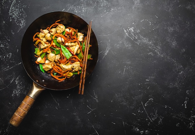 Sauté de poulet aux légumes dans une vieille poêle à wok rustique, baguettes sur fond noir en pierre, gros plan, vue de dessus. Repas traditionnel asiatique/thaï, espace pour le texte