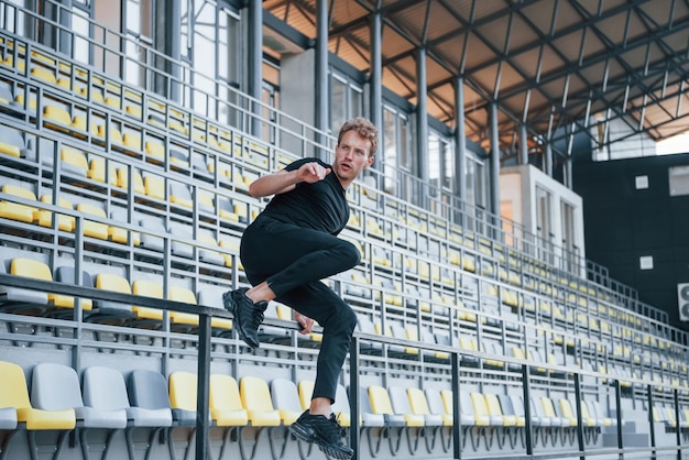 Sauter et faire du parkour sur les gradins Jeune homme sportif en chemise et pantalon noirs à l'extérieur pendant la journée