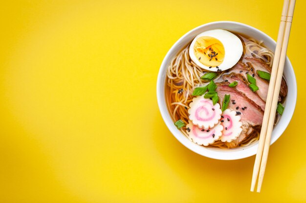 Savoureuse soupe de nouilles japonaises ramen dans un bol en céramique blanche, bouillon de viande, porc tranché, narutomaki, œuf au jaune sur fond jaune pastel. Plat traditionnel du Japon, vue de dessus, gros plan, espace pour le texte