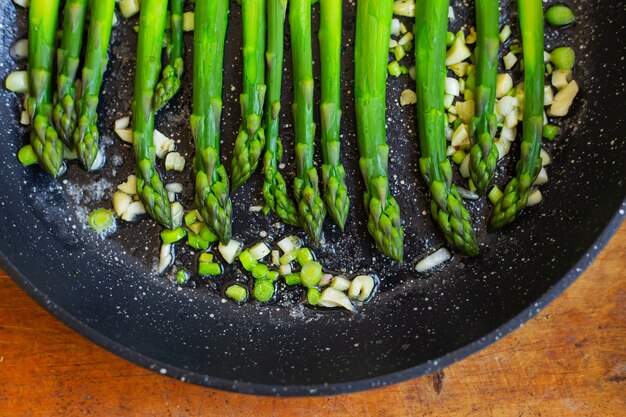 De savoureuses asperges vertes sont frites dans une poêle avec de l'ail, à côté d'une serviette à rayures bleues. Gros plan, vue de dessus.