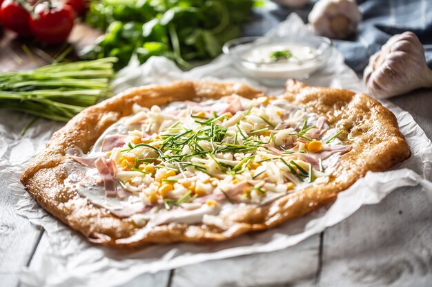 Savoureux langos hongrois servis avec de la crème sure, du jambon, du fromage, des herbes et des légumes en arrière-plan.