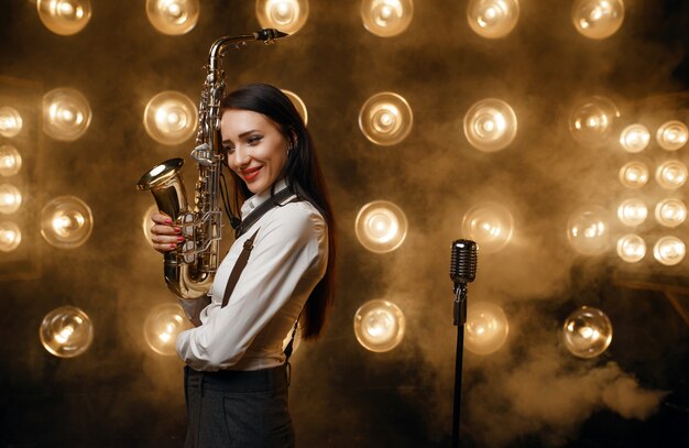 La saxophoniste féminine pose avec le saxophone sur la scène avec des projecteurs. Interprète de jazz jouant sur la scène