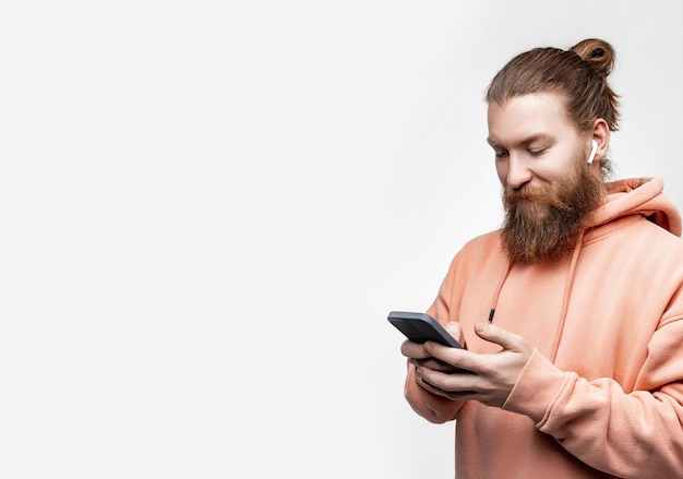 Scandinave bel homme heureux tenant le téléphone et souriant avec un casque sans fil en sweat-shirt orange isolé sur fond gris. Guy avec une coiffure et une barbe au gingembre. Technologies numériques modernes