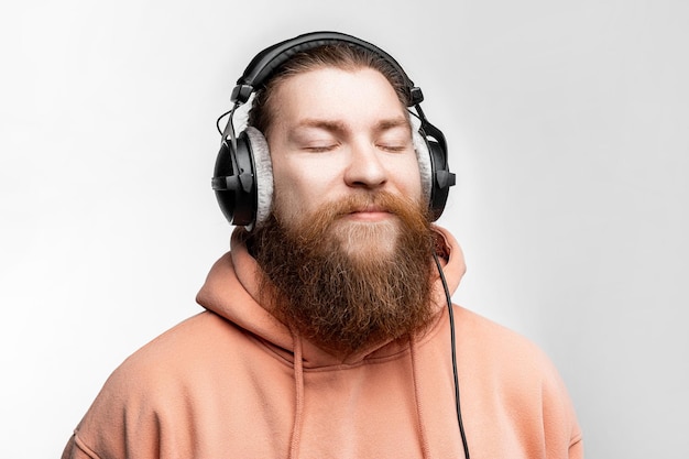 Scandinave bel homme satisfait ferma les yeux et écoute de la musique dans des écouteurs professionnels isolés sur fond gris Heureux mec avec une coiffure et une barbe au gingembre Technologies numériques modernes