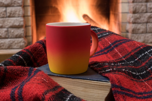 Scène confortable devant la cheminée avec une tasse rouge avec du thé, un livre, un foulard en laine.