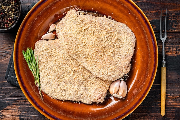 Schnitzel allemand pané cru sur une assiette rustique avec des herbes. Fond en bois sombre. Vue de dessus.