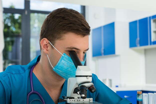 Un scientifique ou un médecin en uniforme avec un phonendoscope sur le cou regarde à travers un microscope et fait des recherches dans un laboratoire. Concept de soins de santé et de biotechnologie