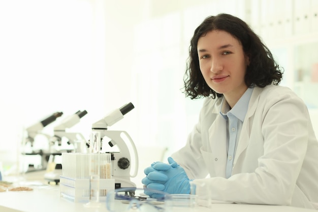 Un scientifique positif en blouse blanche est assis près d'un microscope électronique à table jeune homme dans