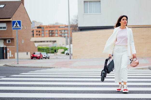 scooter électrique sur la route. Transports écologiques et urbains en ville. Jeune femme souriante traversant la rue avec un scooter électrique à la main