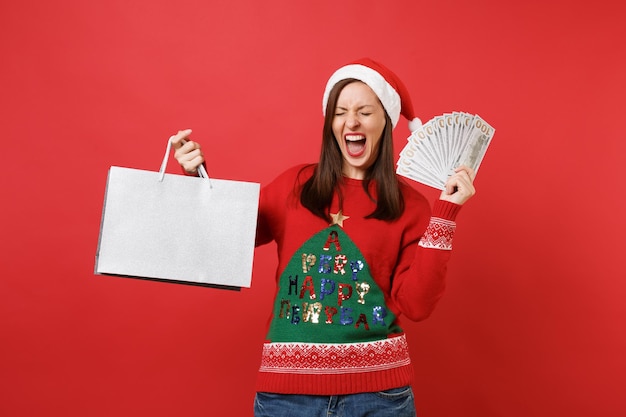 Screaming Santa girl holding beaucoup de billets de banque en dollars, argent liquide, paquets de sacs avec achats après le shopping isolé sur fond rouge. Bonne année 2019 concept de fête de vacances célébration.