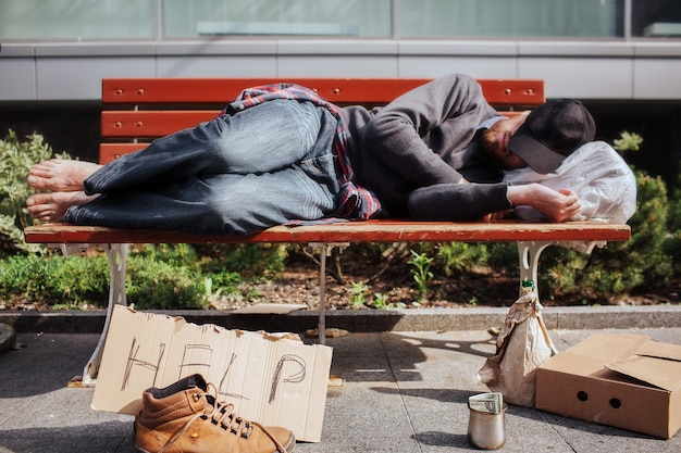 SDF est allongé sur un banc et dort. Hee est fatigué et épuisé. Il y a un sac avec des trucs sous sa tête. Il y a un carton d'aide et une tasse en métal avec de l'argent sur le sol.