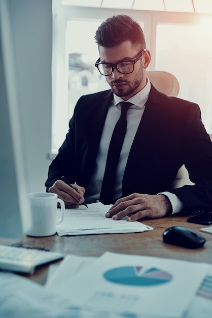 Se concentrer sur le travail. Jeune homme sérieux en tenue de soirée remplissant les formulaires alors qu'il était assis au bureau