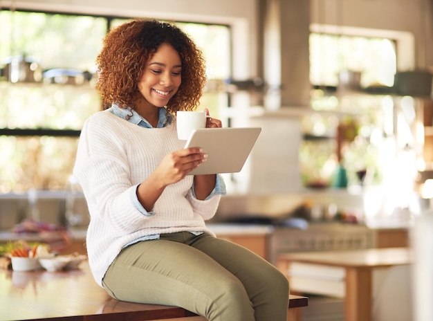 Se détendre n'importe où avec la liberté de la technologie sans fil. Photo d'une jeune femme heureuse utilisant une tablette numérique et buvant du café dans la cuisine à la maison.