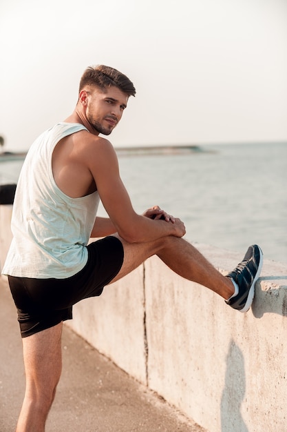 Se préparer à courir. Confiant jeune homme musclé étirant ses jambes avant de courir en se tenant debout à l'extérieur