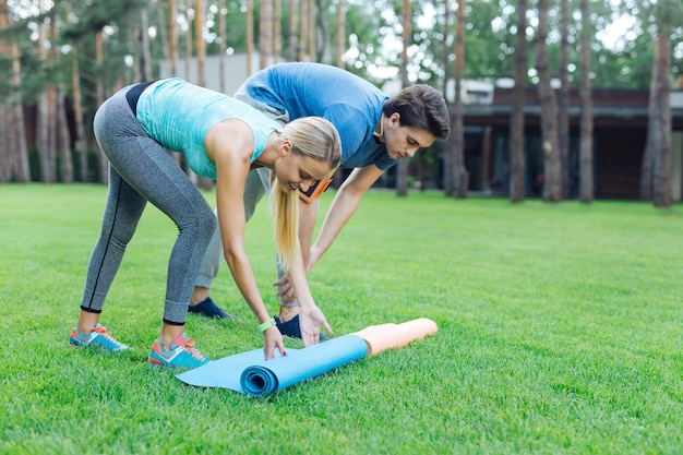 Se préparer pour l'entraînement. Jeunes positifs mettant des tapis de yoga sur l'herbe tout en se préparant pour l'entraînement