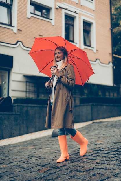 Se promener dans la ville. Toute la longueur d'une jeune femme souriante séduisante en bottes de caoutchouc portant un parapluie et une tasse de café en marchant dans la rue