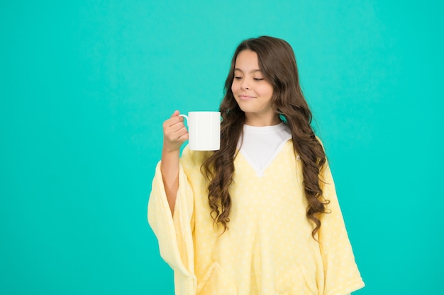 Se sentir à l'aise. Enfant adorable sur fond turquoise. Tenue confortable pour le repos du week-end à la maison. Style confortable. Enfant portant un poncho doux buvant du thé. Notion de bien-être. Petite fille aux cheveux longs.