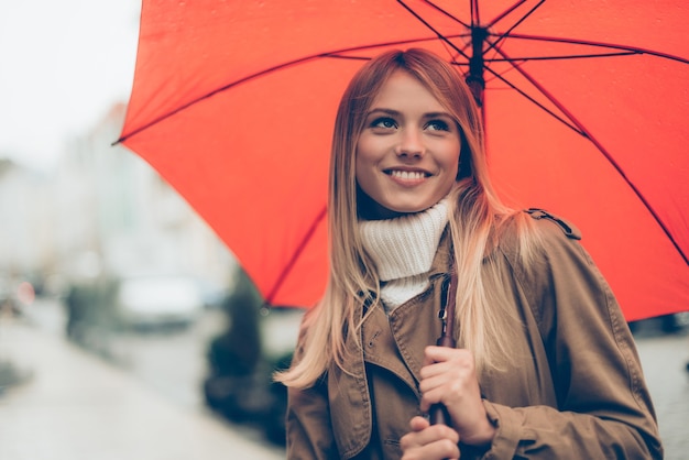 Se sentir au sec et protégé. Jolie jeune femme portant un parapluie et souriant tout en se tenant dans la rue