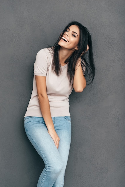 Se sentir bien et avoir fière allure. Jolie jeune femme en tenue décontractée tenant la main dans les cheveux et souriante debout pieds nus sur fond gris