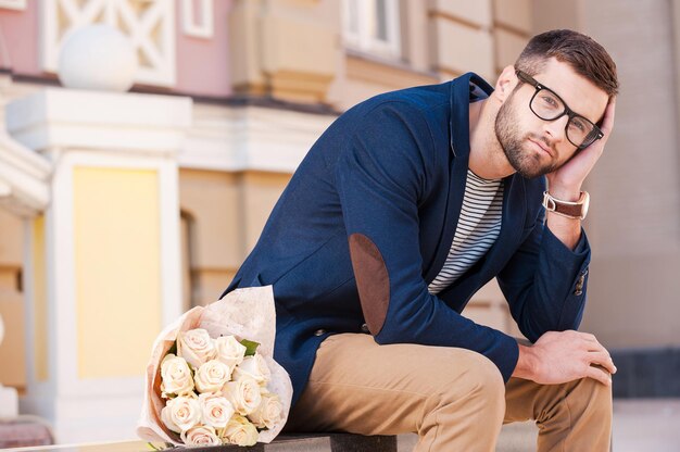 Se sentir désespéré pour elle venir. Jeune homme frustré en veste intelligente s'appuyant la tête sur la main alors qu'il était assis sur le banc avec un bouquet de fleurs posé près de lui