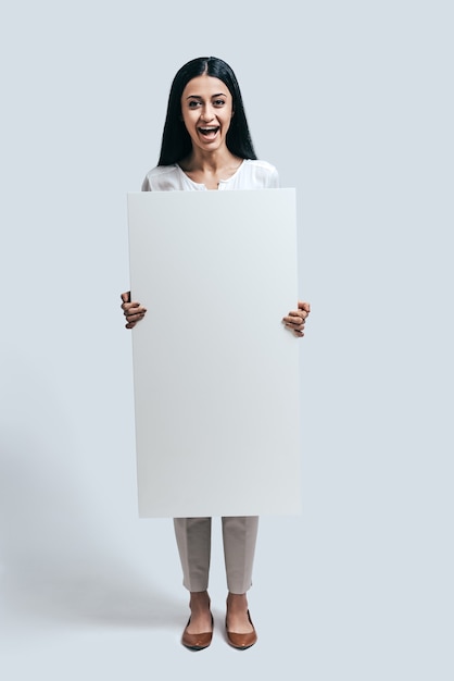 Se sentir excité. Toute la longueur d'une jeune femme confiante tenant un tableau de conférence vierge en se tenant debout sur un fond gris