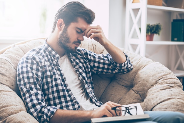 Se sentir fatigué. Jeune homme frustré tenant un livre et se massant le nez alors qu'il était assis sur une grande chaise à la maison et en gardant les yeux fermés