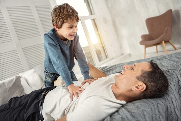 Se sentir heureux. Beau garçon blond joyeux souriant et regardant son papa tandis que son papa allongé sur le dos sur le canapé