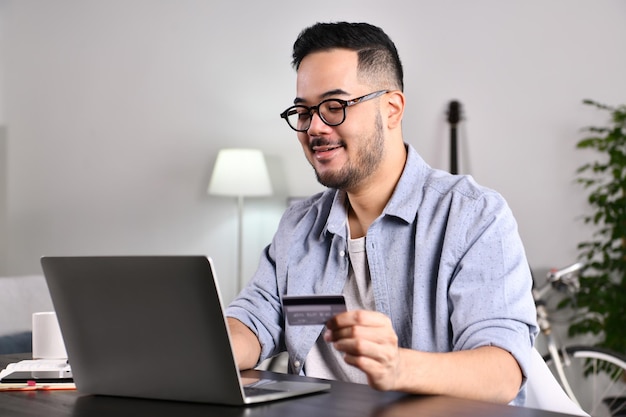 Se sentir heureux et positif. Jeune homme asiatique utilisant un ordinateur portable détenant une carte de crédit pour les achats en ligne et le paiement à la maison.