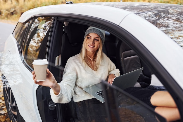 Se trouve à l'intérieur du véhicule avec un ordinateur portable et une tasse de boisson. La fille a un voyage d'automne en voiture. Nouvelle automobile moderne dans la forêt.