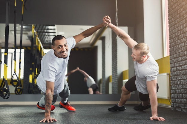 Séance d'entraînement multiethnique d'hommes heureux dans un club de remise en forme. Des gars afro-américains et caucasiens joyeux faisant des exercices de planche ou de pompes en couple, s'entraînant ensemble, espace de copie