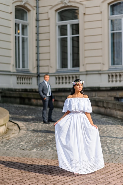 Séance photo de mariage sur le fond de l'ancien bâtiment. Le marié regarde sa mariée poser. Photographie de mariage rustique ou bohème.