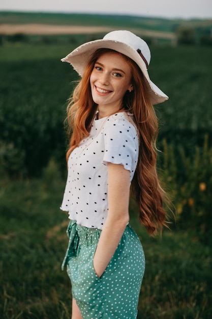 Séance photo en plein air avec une femme au chapeau debout contre un champ vert souriant regardant la caméra moderne...